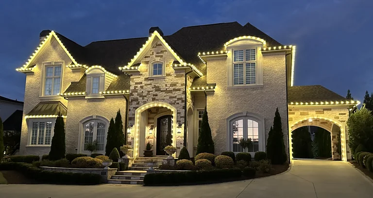 Warm-white Christmas lighting across the roofline of a stone and brick home at dusk.